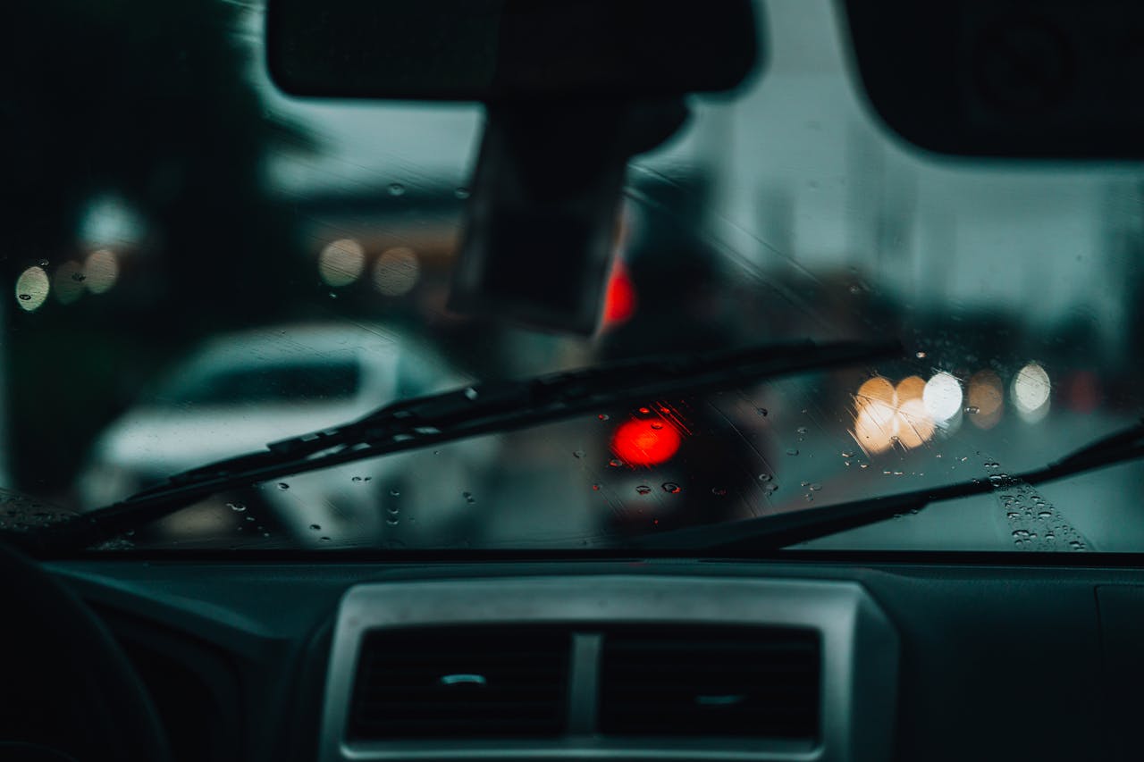 Conventional vs. Beam vs. Hybrid Wiper Blades: Which One Should You Buy? Raindrops on windshield with blurred city lights and cars, creating a moody urban scene.